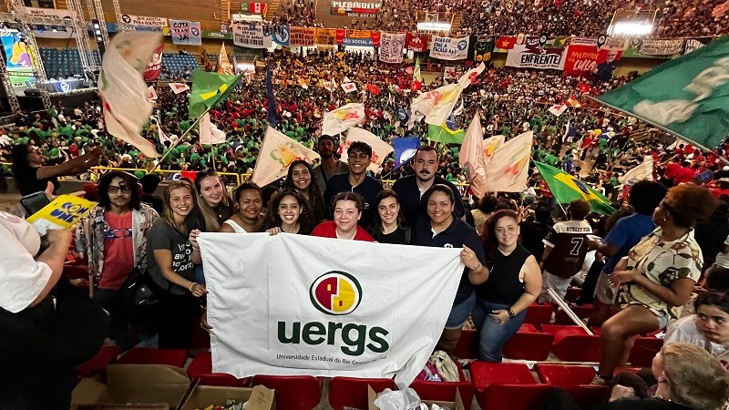 Em um ginásio lotado, um grupo de aproximadamente 15 jovens está em primeiro plano, posando para a foto e segurando uma bandeira branca com o logotipo da Uergs — Universidade Estadual do Rio Grande do Sul. Atrás deles, uma multidão vibra, agita bandeiras e faixas de diferentes cores e movimentos sociais. Algumas faixas ao fundo trazem mensagens. A cena é vibrante, com cores fortes, bandeiras agitadas e um ambiente de participação coletiva e engajamento político.