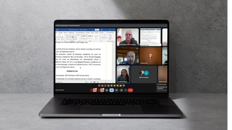 Foto de um notebook aberto sobre uma superfície cinza, exibindo uma videoconferência na tela. A tela está dividida em duas partes principais: do lado esquerdo, um documento de texto (provavelmente uma ata ou pauta de reunião) está aberto no Microsoft Word, com destaque para um trecho de deliberação. Do lado direito, várias janelas de vídeo mostram participantes da reunião online. Algumas câmeras estão ligadas, exibindo rostos de pessoas em ambientes internos, e outras estão desligadas, mostrando apenas nomes ou iniciais. A interface da plataforma usada é o Google Meet. A iluminação geral da cena é neutra e o fundo é liso, em tom cinza-claro.