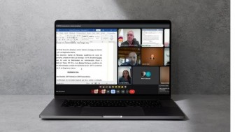  Foto de um notebook aberto sobre uma superfície cinza, exibindo uma videoconferência na tela. A tela está dividida em duas partes principais: do lado esquerdo, um documento de texto (provavelmente uma ata ou pauta de reunião) está aberto no Microsoft Word, com destaque para um trecho de deliberação. Do lado direito, várias janelas de vídeo mostram participantes da reunião online. Algumas câmeras estão ligadas, exibindo rostos de pessoas em ambientes internos, e outras estão desligadas, mostrando apenas nomes ou iniciais. A interface da plataforma usada é o Google Meet. A iluminação geral da cena é neutra e o fundo é liso, em tom cinza-claro.