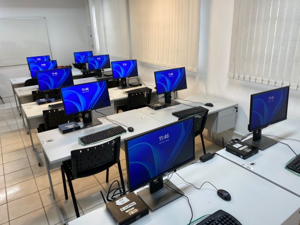 Sala de aula com diversas mesas organizadas em fileiras, cada uma equipada com um kit de computador. Cada kit é composto por um monitor, teclado, mouse e uma unidade central de processamento compacta. Ao todo, há pelo menos dez estações visíveis na imagem. As mesas são brancas, as cadeiras são pretas e têm encosto com pequenos furos quadrados. A sala possui piso claro e paredes brancas, com janelas cobertas por persianas verticais brancas. Todos os monitores estão ligados e exibem a mesma tela azul com a hora “11:46”. Ao fundo, há um quadro branco fixado na parede. A disposição e o ambiente sugerem que o local é utilizado para aulas práticas de informática ou atividades que requerem uso de computadores.
