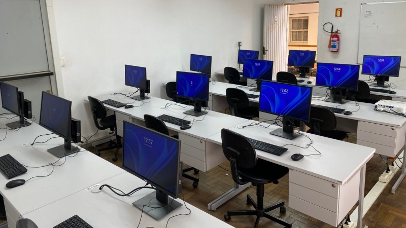 Sala de aula equipada como laboratório de informática, com diversas estações de trabalho organizadas em fileiras. Cada estação conta com monitor, teclado, mouse e CPU compacta, todas ligadas e exibindo uma tela azul com o horário digital — variando entre 12:57 e 13:02. As mesas são brancas com gaveteiros embutidos, e as cadeiras são giratórias, de encosto preto e com rodinhas. O piso é de madeira do tipo parquet.
A sala possui paredes brancas. Ao fundo, vê-se uma porta aberta com grade, uma lousa branca e um extintor de incêndio fixado na parede. Acima da porta, há uma placa de saída de emergência. A iluminação é feita por luminárias fluorescentes fixadas no teto. No canto direito da imagem, um projetor está apoiado sobre uma mesa. O ambiente tem aparência funcional e organizada, preparado para aulas práticas ou capacitações que envolvam uso de computadores.