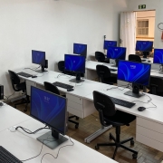 Sala de aula equipada como laboratório de informática, com diversas estações de trabalho organizadas em fileiras. Cada estação conta com monitor, teclado, mouse e CPU compacta, todas ligadas e exibindo uma tela azul com o horário digital — variando entre 12:57 e 13:02. As mesas são brancas com gaveteiros embutidos, e as cadeiras são giratórias, de encosto preto e com rodinhas. O piso é de madeira do tipo parquet.
A sala possui paredes brancas. Ao fundo, vê-se uma porta aberta com grade, uma lousa branca e um extintor de incêndio fixado na parede. Acima da porta, há uma placa de saída de emergência. A iluminação é feita por luminárias fluorescentes fixadas no teto. No canto direito da imagem, um projetor está apoiado sobre uma mesa. O ambiente tem aparência funcional e organizada, preparado para aulas práticas ou capacitações que envolvam uso de computadores.
