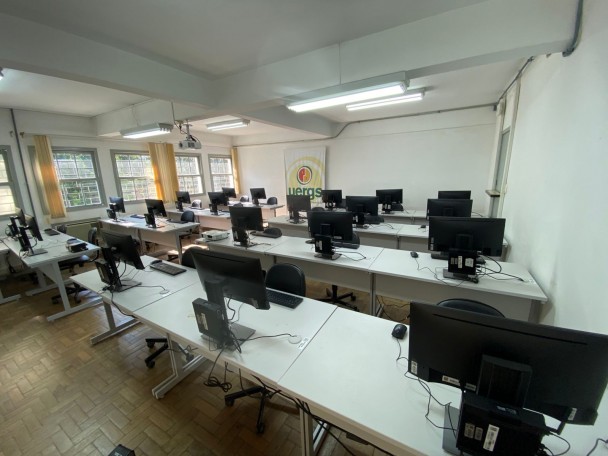 Sala de aula ampla e bem iluminada, equipada com vários computadores organizados sobre mesas brancas distribuídas em fileiras paralelas. Cada estação possui monitor, teclado, mouse e CPU posicionada atrás da tela. Ao fundo, há uma parede branca com um banner da Uergs (Universidade Estadual do Rio Grande do Sul). A sala conta com janelas grandes com cortinas amarelas, por onde entra luz natural. O piso é de parquet de madeira, e o teto tem luminárias fluorescentes. Um projetor está fixado no teto, voltado para a parede do fundo. As cadeiras são pretas, com encosto arredondado. O ambiente transmite um aspecto organizado e propício para aulas práticas ou atividades em grupo com uso de computadores.
