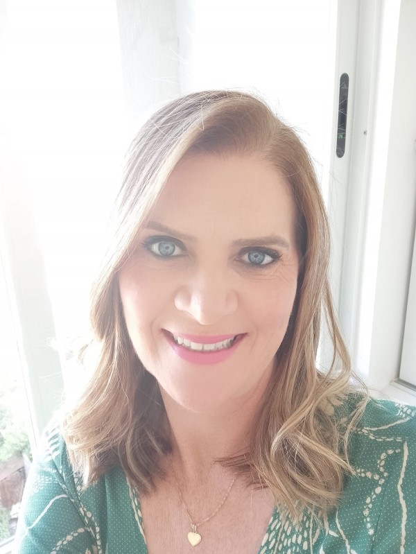Mulher adulta, de pele clara, sorrindo. Tem cabelos loiros, médios e ondulados, repartidos de lado e olhos claros. Veste blusa verde estampada e usa colar com pingente de coração. Ao fundo, uma janela com luz natural.