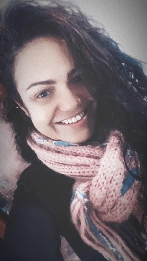 Mulher adulta sorrindo. Tem cabelos escuros, longos e cacheados, pele clara e sobrancelhas marcadas. Usa um cachecol de tricô em tom rosado e roupa escura. A imagem tem iluminação suave e fundo neutro.