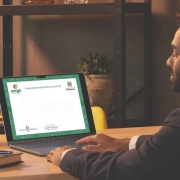 Imagem em ambiente interno, com ilumina&ccedil;&atilde;o suave. &Agrave; direita, um homem jovem adulto, de pele clara, cabelos curtos escuros e barba aparada, veste terno escuro e camisa clara. Ele est&aacute; sentado de perfil em frente a uma mesa de madeira, olhando para a tela de um notebook aberto.

Na tela do computador aparece um diploma digital da Universidade Estadual do Rio Grande do Sul (Uergs). No topo do documento est&atilde;o vis&iacute;veis o logotipo da Uergs, o nome &ldquo;Universidade Estadual do Rio Grande do Sul&rdquo; e o bras&atilde;o do Governo do Estado. O diploma possui moldura verde e espa&ccedil;o central claro para as informa&ccedil;&otilde;es do documento.

Sobre a mesa, &agrave; esquerda do notebook, h&aacute; uma lumin&aacute;ria preta, um caderno com capa decorada, um bloco de notas e uma caneta azul. Ao fundo, uma estante escura com prateleiras cont&eacute;m livros e um vaso com planta.