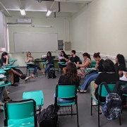 Sala de aula com paredes claras e ilumina&ccedil;&atilde;o fluorescente no teto. &Agrave; esquerda, h&aacute; janelas amplas com grades, por onde entra luz natural. No fundo da sala, um quadro branco ocupa a parede, com um aparelho de ar-condicionado instalado acima.

Cerca de 15 pessoas, entre jovens e adultos, est&atilde;o sentadas em cadeiras escolares com prancheta, organizadas em c&iacute;rculo. A maioria segura folhas de papel e parece acompanhar uma leitura ou discuss&atilde;o. No centro do c&iacute;rculo, duas mulheres est&atilde;o com maior destaque: uma fala com gestos das m&atilde;os, enquanto a outra observa um material.

Os participantes vestem roupas casuais, como camisetas, cal&ccedil;as jeans e t&ecirc;nis. Algumas mochilas est&atilde;o apoiadas no ch&atilde;o ao lado das cadeiras.