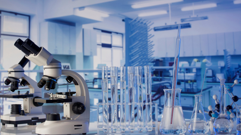 &Agrave; esquerda, doi microsc&oacute;pios sobre bancada. &Agrave; direita, v&aacute;rios tipos de tubos de ensaio e frascos de vidro enfileirados. Ao fundo, ambiente de laborat&oacute;rio, com outros equipamentos.