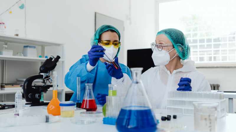Duas mulheres est&atilde;o em um laborat&oacute;rio, manipulando tubo de ensaio com um l&iacute;quido vermelho. Elas usam equipamentos de prote&ccedil;&atilde;o e seguran&ccedil;a, como toucas, &oacute;culos, m&aacute;scaras, luvas e jalecos. 